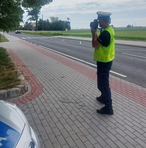 Policjant wykonuje pomiar prędkości pojazdu w rejonie przejścia dla pieszych. Z lewej strony na poboczu zaparkowany policyjny radiowóz.