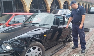 Teren parkingu strzeżonego. Policjant stoi przy skradziony pojeździe, który jest uszkodzony.