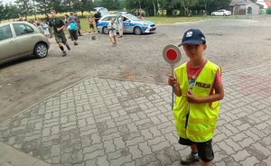 Chłopiec w żółtej kamizelce z napisem policja i czapką koloru granatowego na łowie. w ręku trzyma tarcze do zatrzymywania pojazdów. W oddali policyjny radiowóz.