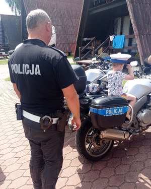 Policjant ruchu drogowego stoi przy motocyklu służbowym. Na nim siedzi mały chłopiec. na głowie ma ubraną białą czapkę symbolizującą służbę ruchu drogowego.