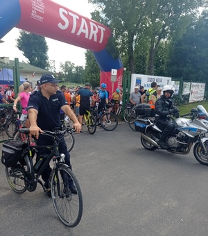 Miejsce startu rajdu. Z lewej strony przy bramie stoi dzielnicowy z rowerem, z prawej strony bramy stoi policjant ruchu drogowego na motocyklu.