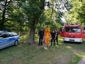 Przedstawiciele służb, z lewej strony policyjny radiowóz, po środku stoi dzielnicowy, tygrysek maskotka, oraz dwóch strażaków. Z prawej strony fotografii wóz strażacki.