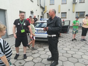 Policjant ruchu drogowego rozmawia z opiekunem obozu. Za nimi widoczne dzieci, które oglądają radiowóz.