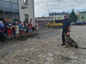 Przewodnik psa opowiada o pracy z czworonogiem. Z lewej strony dzieci ustawione wzdłuż budynku.