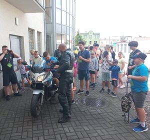 Policjant ruchu drogowego pokazuje, gdzie na motocyklu służbowym włączyć sygnały uprzywilejowania. Wokół niego skupione dzieci.