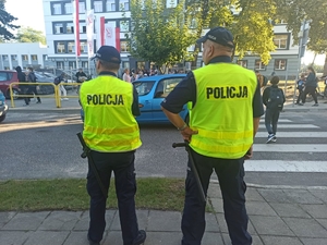 dwóch  umundurowanych policjantów stoi przed budynkiem szkoły, gdzie przemieszczają się dzieci