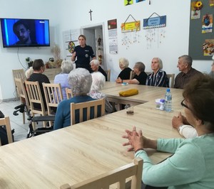 seniorzy siedzą przy stole, przed nimi stoi policjantka