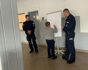 uczeń wykonuje rysunek na tablicy, obok stoi dwóch umundurowanych policjantów