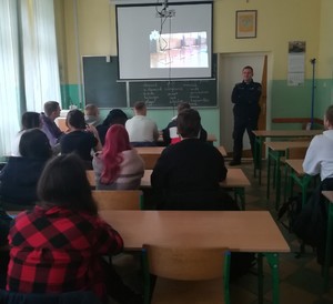 policjant stoi przed uczniami, którzy siedzą w szkolnych  ławkach