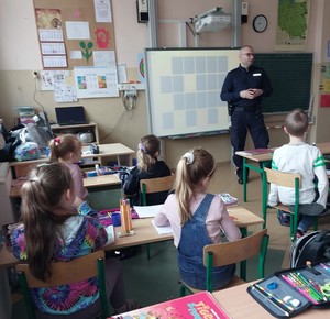 policjant  stoi w  klasie lekcyjne, a w ławkach siedzą dzieci