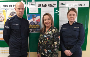 policjantka, policjant oraz kobieta w ubraniu cywilnym stoją przy tablicy informacyjnej, gdzie znajduje się miedzy innymi plakat zachęcający do wstąpienia  w szeregi Policji