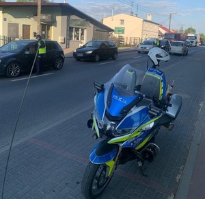 na  pierwszym planie stoi policyjny motocykl, a dalej policjantka  bada kierowcę osobówki