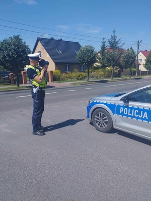 Policjant ruchu drogowego ubrany w żółtą kamizelkę odblaskową wykonuje pomiar prędkości nadjeżdżających pojazdów. Przed nim na poboczu drogi stoi zaparkowany radiowóz.