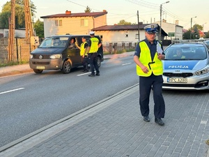 Policjanci podczas działań. Z lewej strony funkcjonariusze sprawdzają trzeźwość kierującego busem. Z prawej strony zaparkowany radiowóz policyjny. W środku na miejscu pasażera siedzi policjant. Do radiowozu zmierza inny policjant, który trzyma w ręku teczkę.