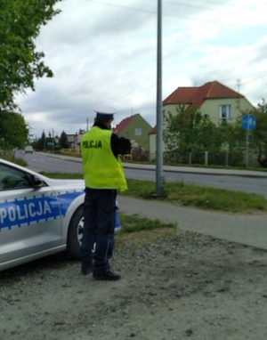 Policjant obserwuje ruch drogowy. Z lewej strony zaparkowany radiowóz, a z jego prawej strony stoi policjant. Przed nim przebiega droga.