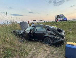 Pobocze drogi S5. w rowie leży uszkodzony pojazd koloru ciemnego. Z lewej strony strony siatka odgradzająca od drogi S5. za uszkodzonym pojazdem policjantka, dwóch strażaków i kobieta. z prawej strony samochodu stoją dwie walizki. Z prawej strony do góry na pasie ruchu stoi karetka pogotowia.