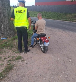 Na poboczu drogi znajduje się policjant i mężczyzna siedzący na motocyklu. obok przebiega droga.