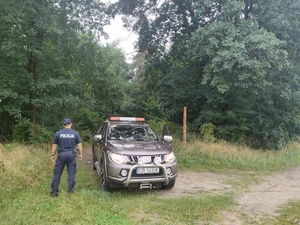 Teren leśny. Z lewej strony tyłem stoi policjant. Z prawej strony samochód Państwowej Straży Łowieckiej.