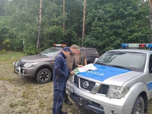 Teren leśny. Zaparkowany pojazd Straży Łowieckiej oraz Policji. Na masce policyjnego radiowozu rozłożona mapa. Dwaj strażnicy i policjant analizują ją.