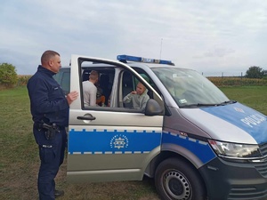 Z lewej strony stroi policjant, którzy trzyma rękę na drzwiach radiowozu. W środku znajdują się dzieci.