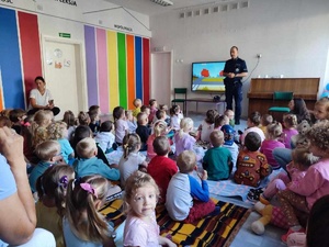 Na środku sali referuje policjant. Przed nim na dywanie siedzą dzieci. Za jego plecami wyświetlana jest prezentacja multimedialna.