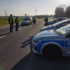 Z prawej strony fotografii policyjne pojazdy zaparkowane obok siebie. Na jezdni policjanci ruchu drogowego, którzy wykonują sprawdzenia stanu trzeźwości oraz nadjeżdżające od lewej strony pojazdy.