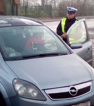 Policjant podczas kontroli pojazdu. Funkcjonariusz stoi z prawej strony, z lewej strony pojazd osobowy, który ma otwarte drzwi od strony kierującego.