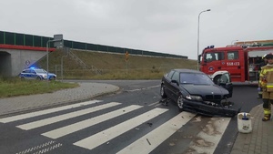 Miejsce zdarzenia drogowego. Na pierwszym planie przejście dla pieszych. Z prawej strony na drodze znajduje się uszkodzony samochód marki BMW, za nim pojazd straży pożarnej oraz strażacy. Z lewej strony na drodze dojazdowej zaparkowany pojazd policyjny z włączonym niebieskim światłem ostrzegawczym. Za radiowozem przebiega droga ekspresowa S5.