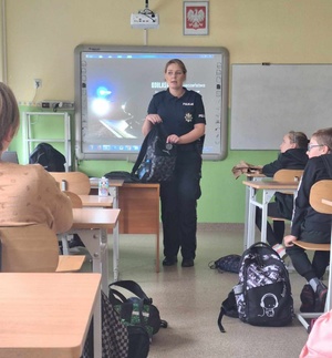 Policjantka podczas prelekcji. Za nią prezentacja multimedialna do akcji &quot;Świeć przykładem&quot;. Policjantka trzyma w ręku plecak i pokazuje element odblaskowy. przed nia dzieci w ławkach.