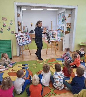 Policjantka podczas prelekcji. W ręku trzyma tubę profilaktyczną. Przed nią na dywanie siedzą dzieci.