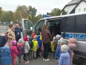 Dzieci oglądają policyjny radiowóz. Przedszkolaki ustawione w dwie kolejki po kolei podchodzą do samochodu.  Z lewej strony przy drzwiach kierowcy stoi policjant.