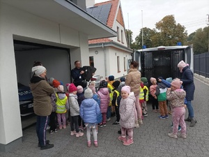 Przed budynkiem komisariatu policjant pokazuje kamizelkę kuloodporną. Za nim w garażu zaparkowany policyjny radiowóz. Z prawej strony na podjeździe stoi drugi samochód marki Fiat Ducato z otwartą częścią służącą do przewozu osób zatrzymanych.