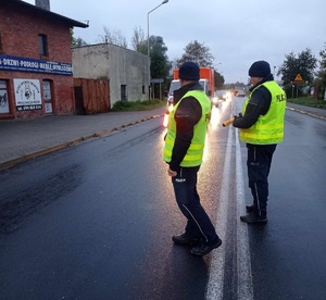 Policjanci ruchu drogowego ubrani w żółte kamizelki stoją na środku jezdni. W tle budynki.