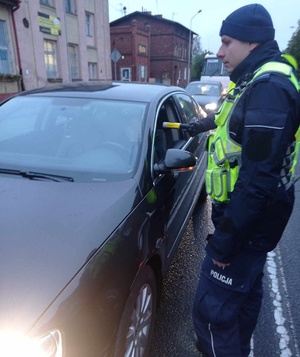 Policjanci drogówki alkomatem sprawdzają trzeźwość kierowcy w granatowym samochodzie osobowym. W tle budynki.