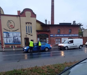 Policjanci ruchu drogowego kontrolują kierowcę w niebieskim samochodzie osobowym. Za nim stoi biała osobówka.