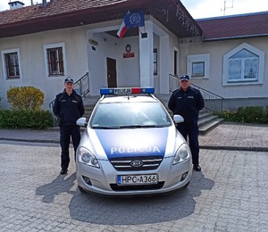 mł. asp. Mateusz Przygoda i st. asp. Krzysztof Zientek. Pomiędzy nimi policyjny radiowóz, a za nimi budynek Komisariatu Policji w Barcinie