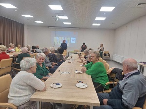 Policjantka stoi przy tablicy multimedialnej. Przed nią przy stołach siedzą seniorzy.