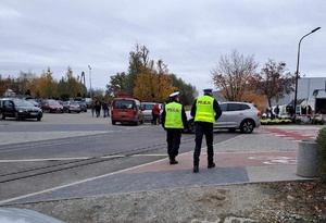 Policjanci ruchu drogowego w rejonie cmentarza obserwują ruch drogowy. W oddali cmentarz i parking samochodowy.