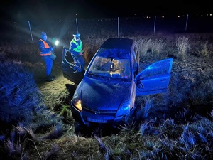 Policjant ruchu drogowego zagląda do wnętrza uszkodzonego samochodu osobowego, który brał udział w zdarzeniu drogowym. Pojazd ma widoczne uszkodzenia. Przy policjancie stoi mężczyzna zajmujący się obsługą drogi S5, który świeci latarką. Wokół panuje mrok.