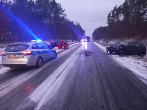 Droga w miejscu zdarzenia. Na pierwszym planie policyjny radiowóz. Przed nim uszkodzone samochody marki Renault i Seat po obydwu stronach drogi. W oddali straż pożarna.