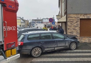 Z lewej strony widoczny fragment pojazdu straży pożarnej. Dalej uszkodzone dwa samochody osobowe znajdujące się przy budynku.