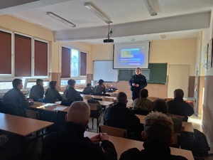 Zajęcia w klasie mundurowej. Policjantka stoi na środku sali. Za nią rzutnik multimedialny. Przed nią uczniowie w ławkach.