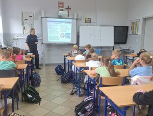 policjant a stoi przed dziećmi  siedzącymi w  ławkach